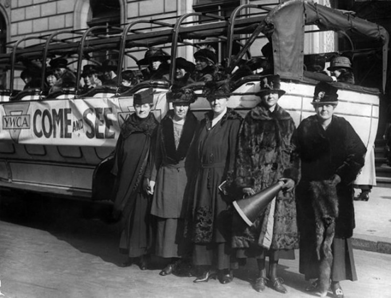 In this vintage black and white photo, 5 YWCA women advocates stand in front of a bus full of more women. A banner labeled 'YWCA Come and See' is attached to the side of the bus.