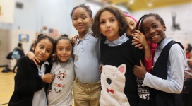 Six elementary school girls of varying height stand side-by-side as they lean on and hug each other while smiling into the camera.