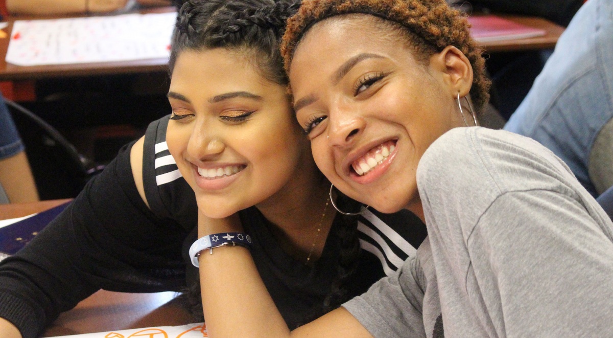 This photo is a close up of two teen women sitting at a desk and smiling into the camera with their heads against each others.