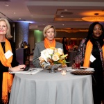Three women stand around a small round table at a professional networking event. They all wear orange drapes across their shoulders. These women are YWCA NYC AWL 2018 Honorees Charlene Eigenberg, Beth Ann Bovino & Camille Joseph-Goldman