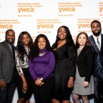 In this photo, 8 people stand in a row before a white step and repeat featuring the YW orange logo. Pictured are YW Staff from left to right: YWCA NYC CEO Rosemarie Bonelli, Senior Director of Out-Of-School Time Programs Phillip Colas, Program Director Ewuraesi Dadzie, Program Director Janet Cooper, Program Director Karlene James, Benefits Administrator Nicole Doherty, Program Director Christopher Anderson