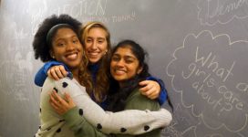 Three teenaged women embrace while smiling into the camera. Girl empowerment notes are written on the chalkboard wall behind them.