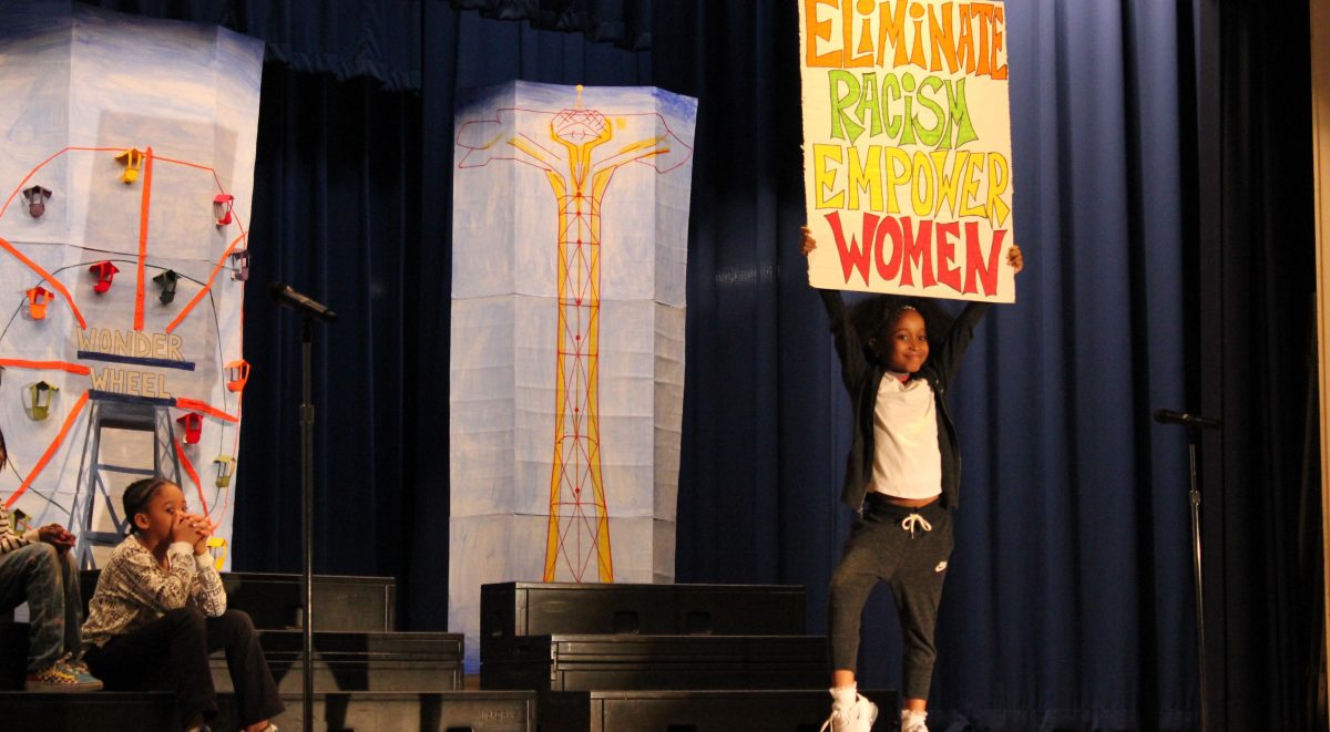PS 9o students take a Stand Against Racism at the annual event. In this photo an elementary school girl poses on stage holding a sign that reads Eliminate Racism. Empower Women.