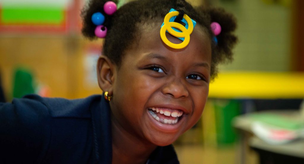 Young girl from our Out of School Time Program smiles brightly into the camera while sitting in a classroom.